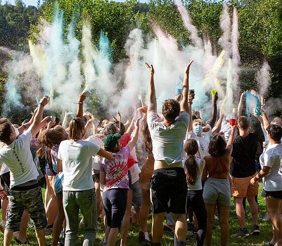 Eine große Gruppe Jugendlicher wirft bei ausgelassener Stimmung bunte Farbpulver in die Luft auf einer sonnigen Wiese.