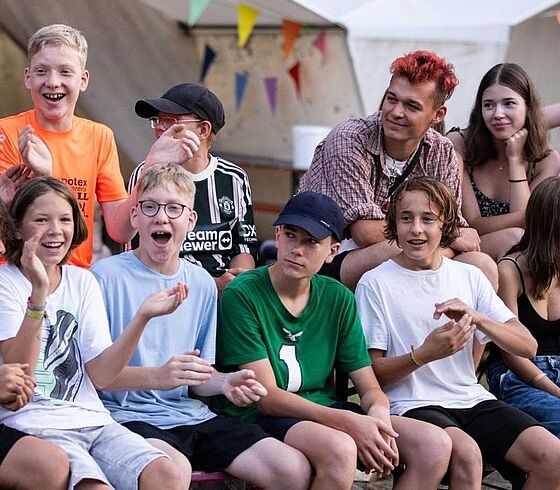 Eine fröhliche Gruppe Jugendlicher sitzt vor Zelten und lacht ausgelassen in die Kamera.