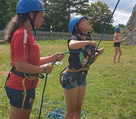 Zwei junge Personen mit Klettergurten und Helmen sichern eine weitere Person, die an einer hohen, steinernen Felswand im Donautal klettert. Der Boden ist mit Gras bedeckt, im Hintergrund sind Bäume und ein Geländer zu sehen.